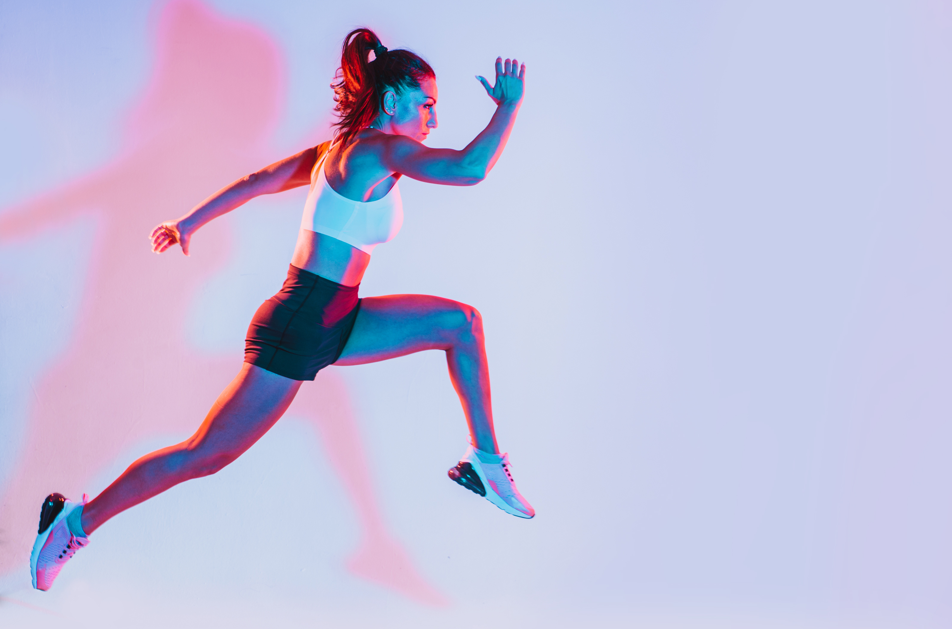 Sportlerin in weißem Sporttop und schwarzer Shorts springt in einer dynamischen Laufbewegung vor hellem Hintergrund mit blau-rosa Lichteffekten.